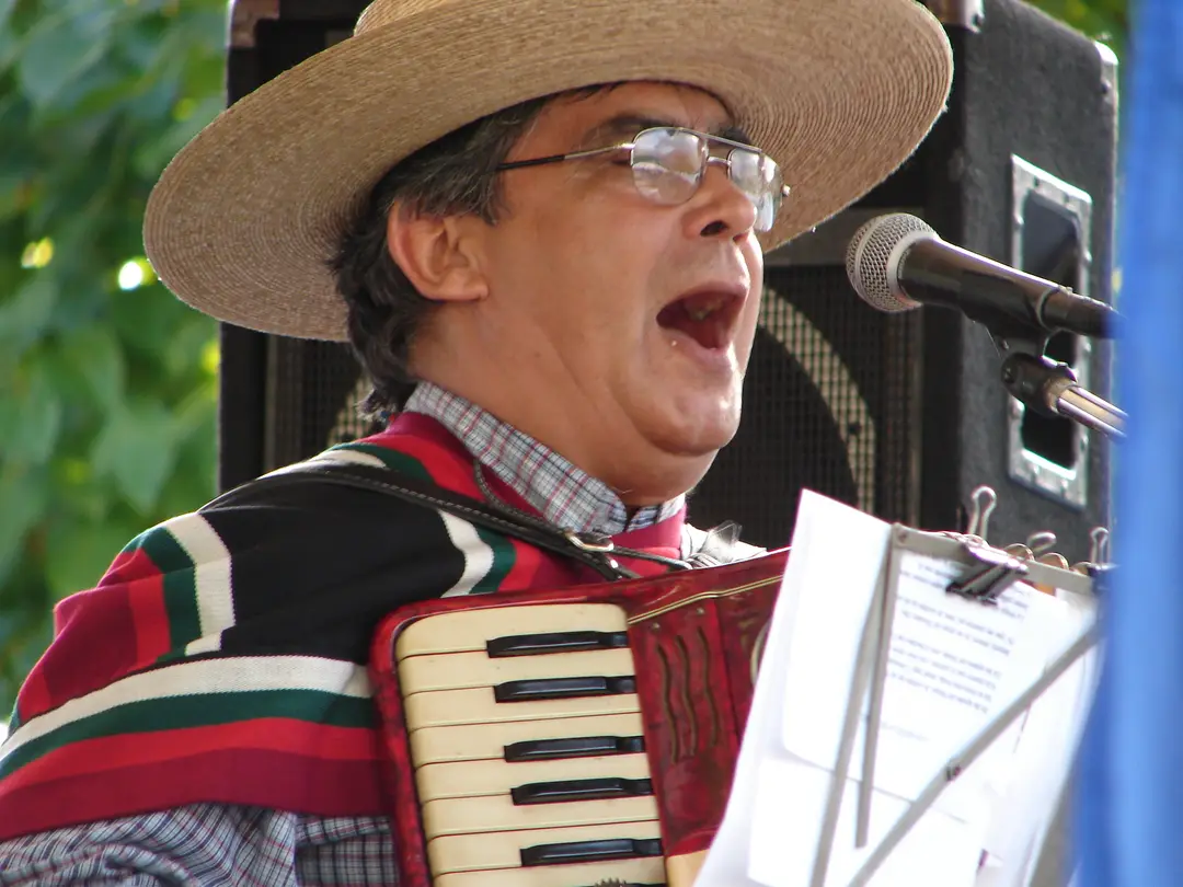 Músico con chupalla y manta cantando y tocando acordeón durante una celebración de Fiestas Patrias.